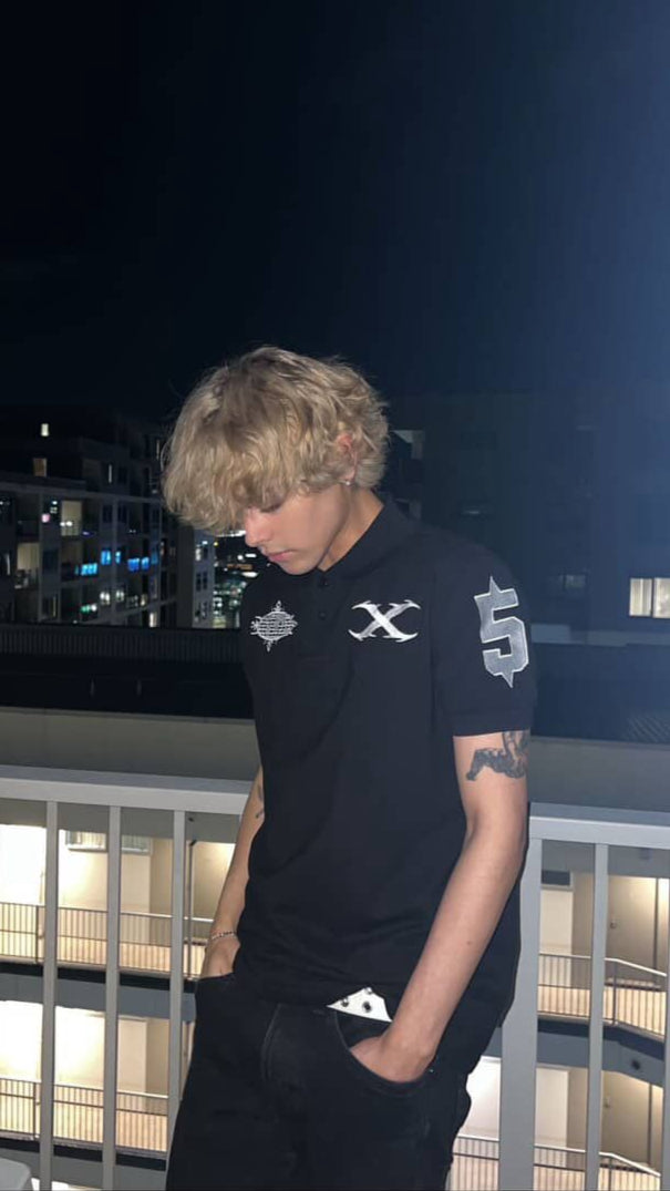 Person wearing a black polo-shirt with white designs, standing on a balcony at night.